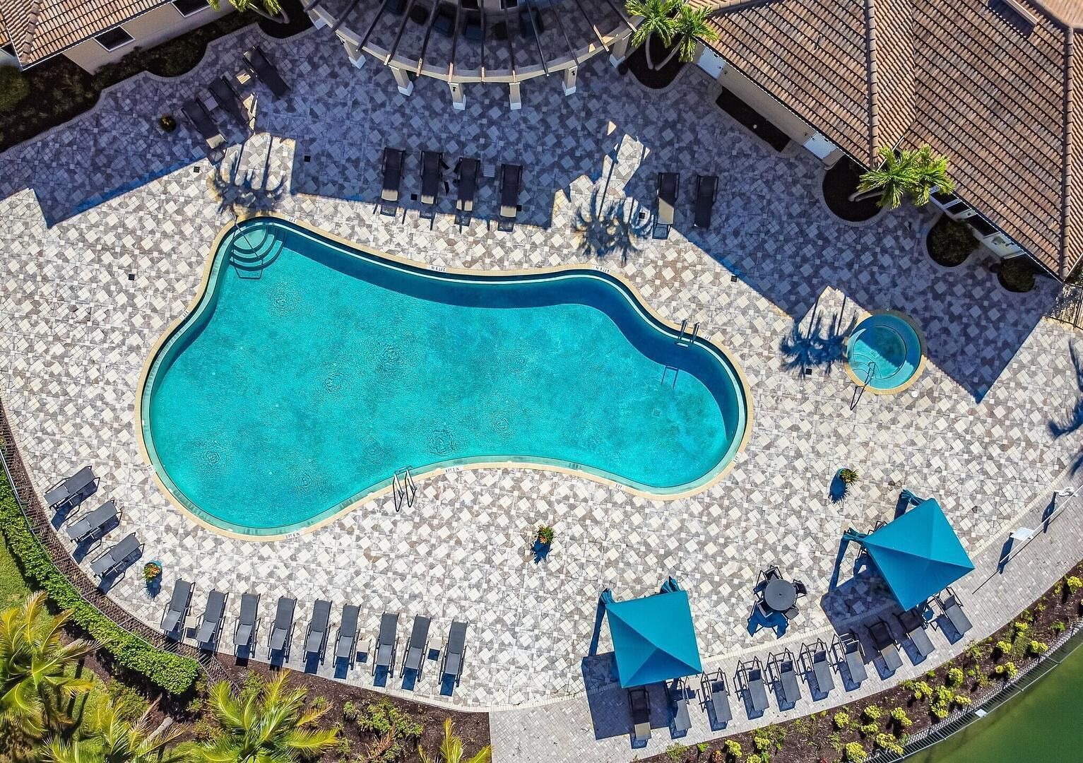 An aerial view of a swimming pool surrounded by lounge chairs and umbrellas, with a tiled deck and palm trees.
