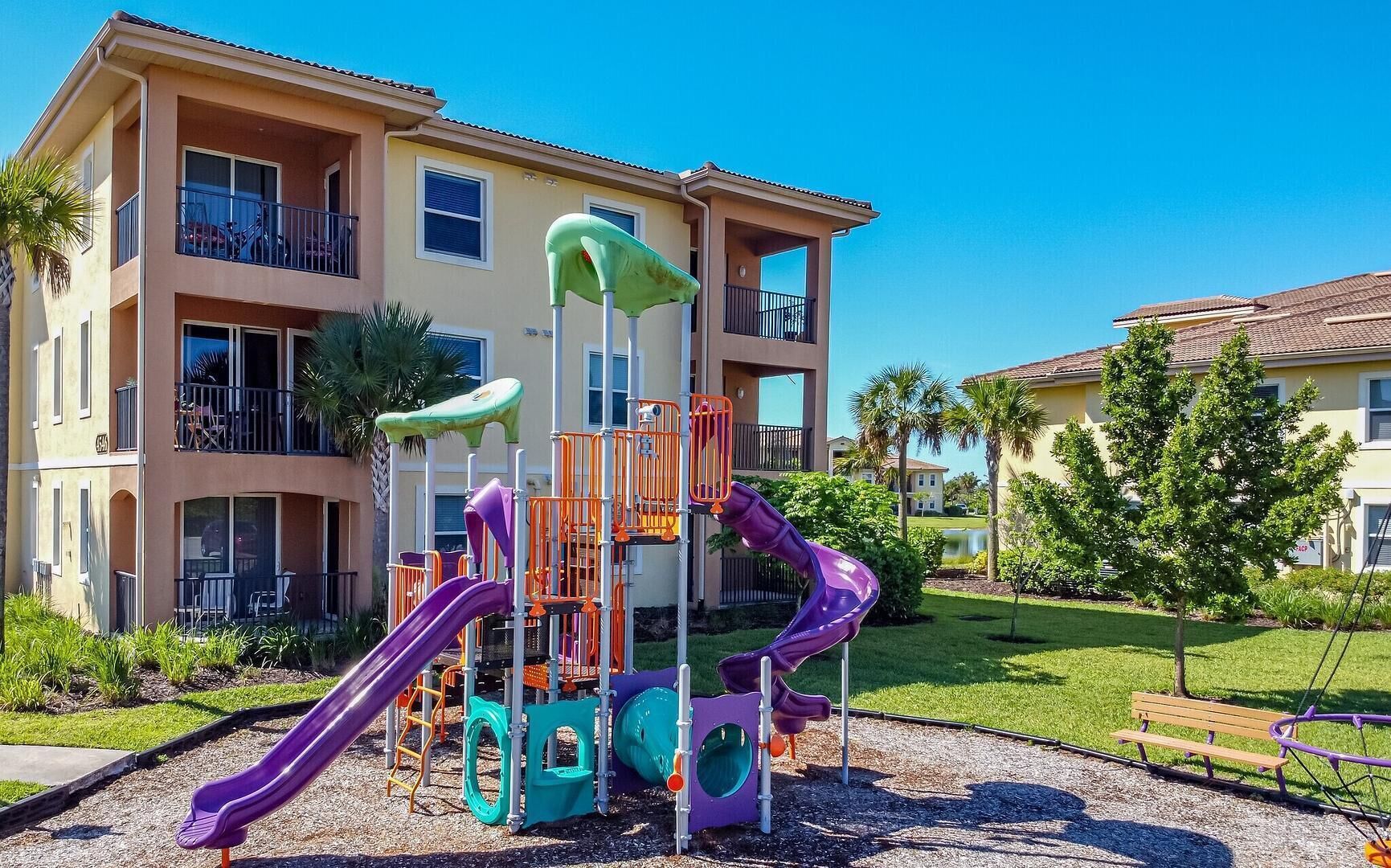 A playground with slides and climbing structures in front of a multi-story building with balconies.