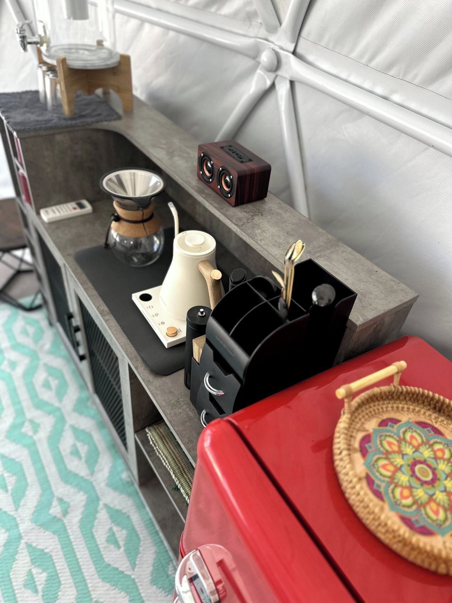 A kitchenette with a coffee maker, a kettle, a speaker, and a red suitcase.