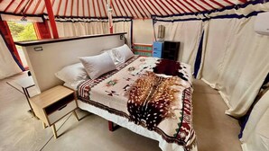 A bed with a patterned blanket, a small bedside table, and a window with curtains. - Roadside Mongolian Yurt (Teasdale)