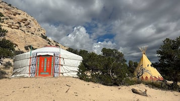 A yurt with a red door and a traditional yellow tipi nearby, set against a backdrop of a rocky hill and trees under a cloudy sky.