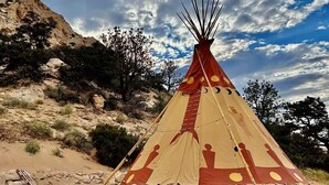 A traditional tipi with a conical shape and a wooden top, set against a rocky landscape with sparse vegetation. - Native American Bison Tipi (Teasdale)
