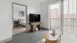A modern hotel room with a large TV on a wooden stand, a bed with patterned pillows, a desk with a chair, and a view of a red building through the balcony. - Landing | Stylish 1BD, Gym, Clubhouse (Richmond)