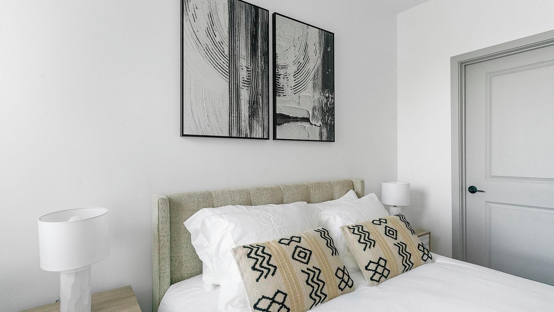 Un dormitorio ordenado con una cama, dos obras de arte colgadas en la pared, una lámpara de noche y una puerta.