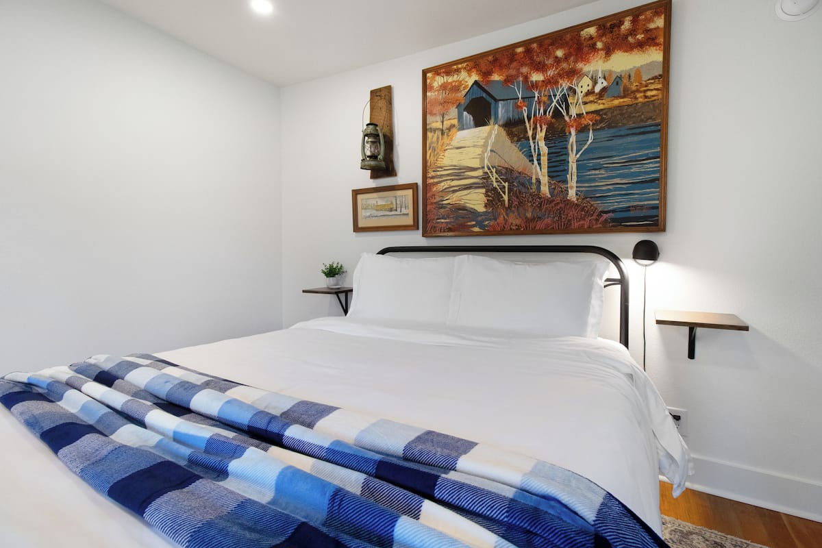 A neatly made bed with a blue and white checkered blanket, a painting of a landscape on the wall, and a small plant on a bedside table.