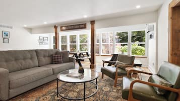 A spacious living room with a gray sofa, a glass coffee table, and leather armchairs. There are large windows providing natural light and a view of greenery outside.