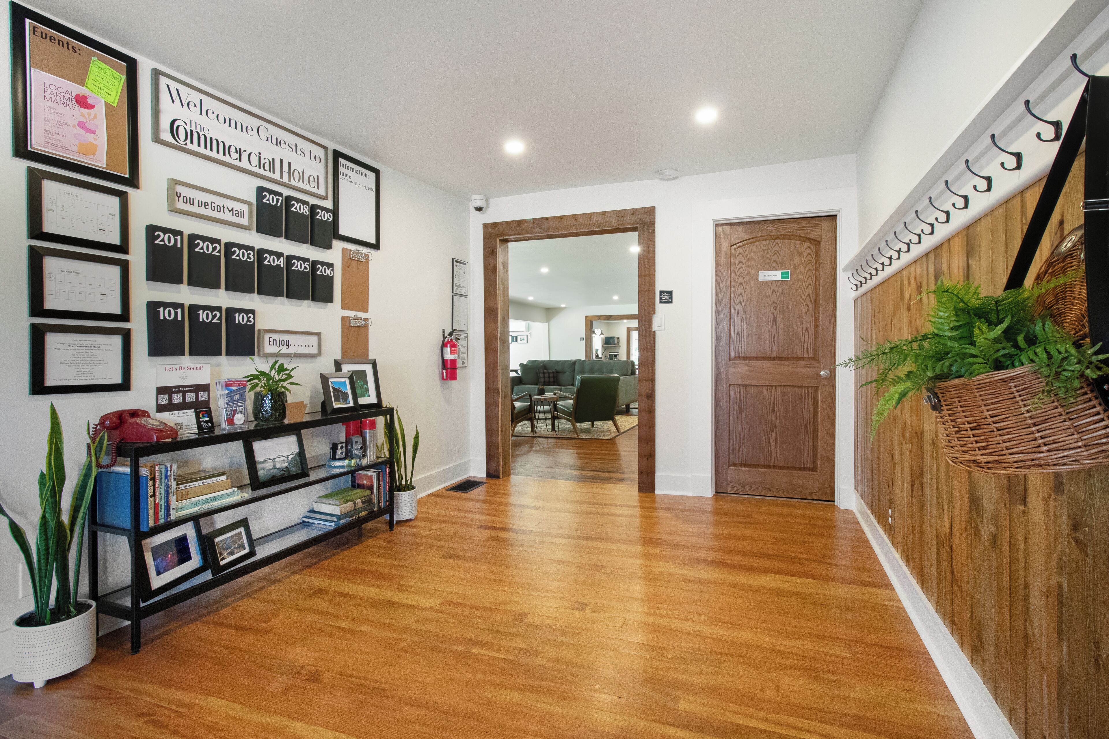A hotel hallway with a wooden floor, a reception desk with framed certificates, a bookshelf, and a wall-mounted sign indicating room numbers.