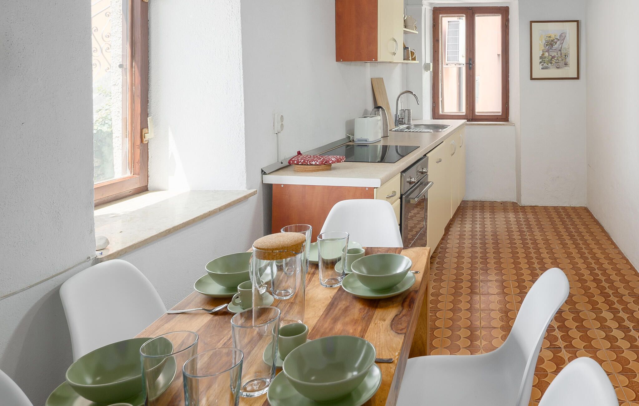 Une cuisine compacte avec un coin repas, comprenant une table en bois dressée avec de l’assiette et des verres verts, un évier et une fenêtre donnant sur un paysage agréable.