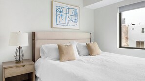 A bedroom with a bed, a nightstand, a lamp, and a window with a view of a building. - Landing | Spectacular 2BD, Gym (Philadelphia)