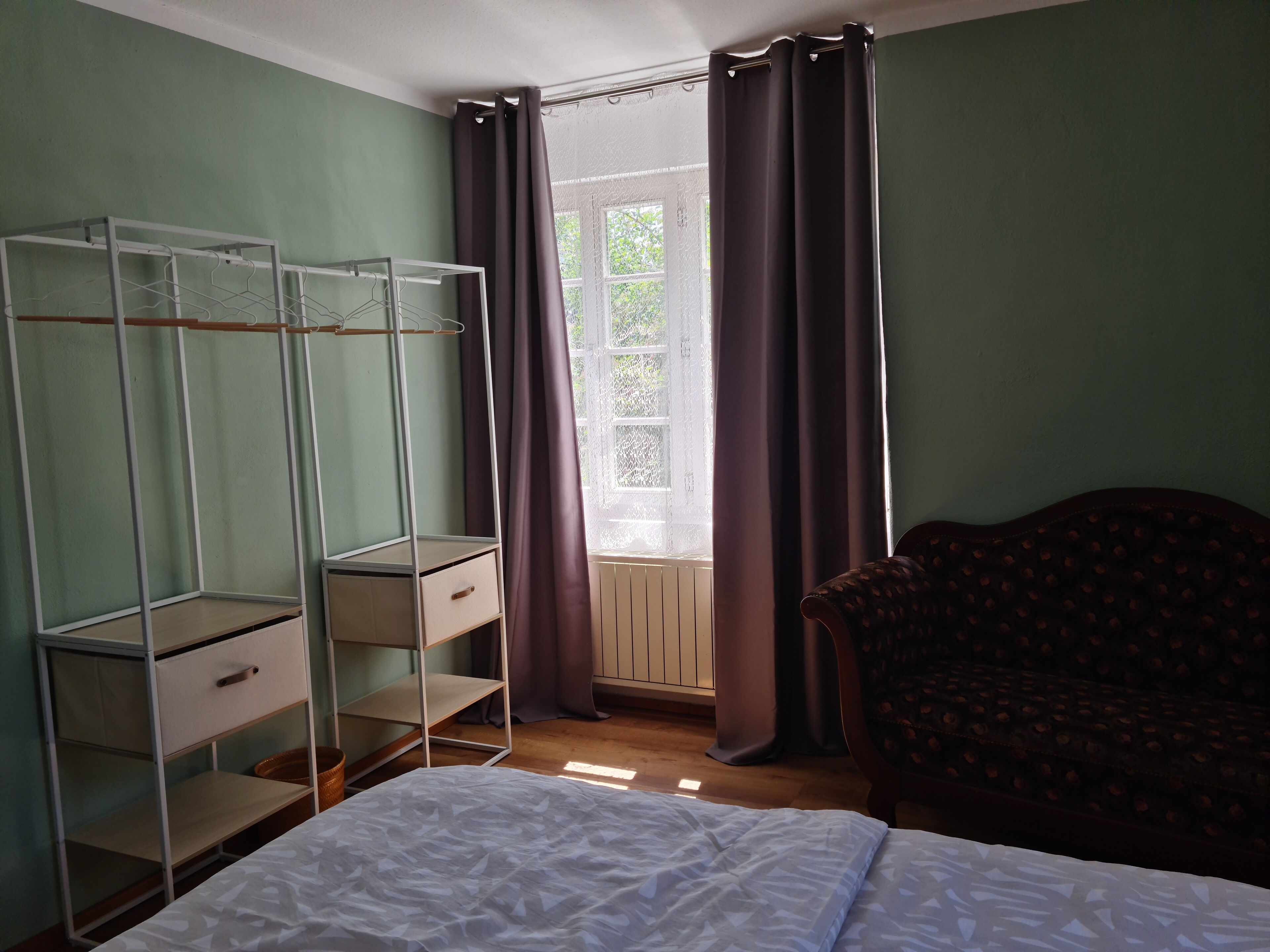 Ein Schlafzimmer mit Bett, einem Kleiderschrank mit vielen Ablagen, einem Sessel und einem Fenster mit Vorhängen.