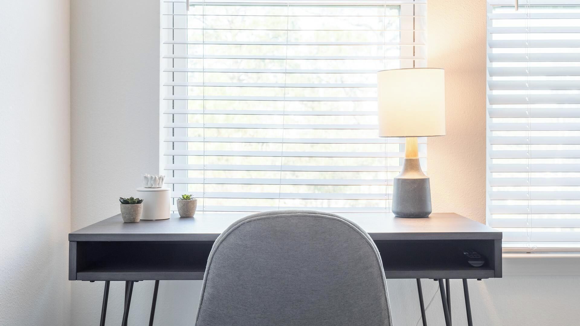 A modern desk with a lamp, two small potted plants, and a chair in front of a window with blinds.