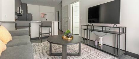 Un salon moderne avec un canapé gris, une table basse ronde agrémentée d’objets décoratifs, un téléviseur à écran plat fixé sur un meuble TV noir, et une cuisine en arrière-plan.