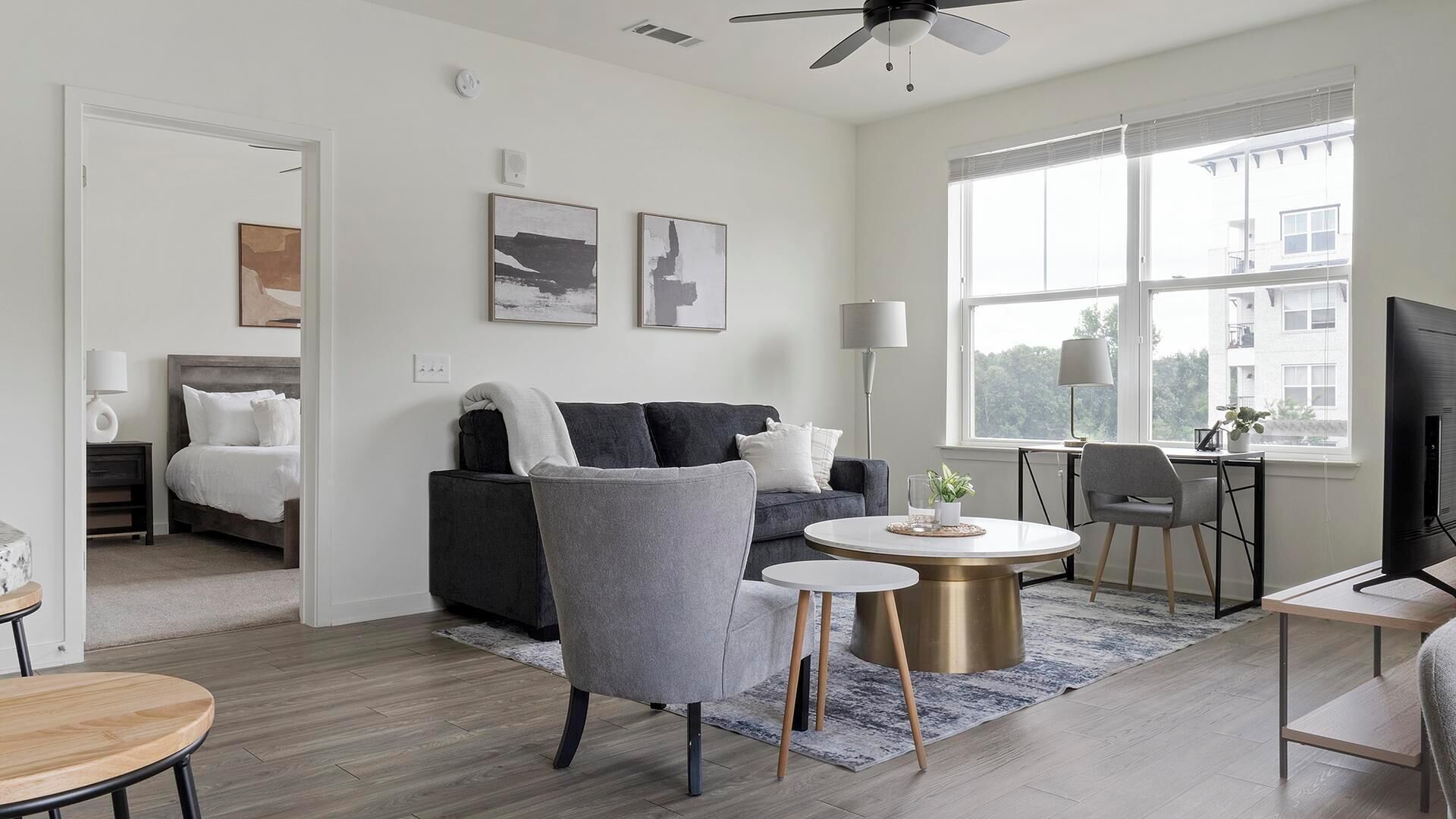 Ein modernes Wohnzimmer mit einem grauen Sofa, einem runden Couchtisch und einem Fernseher. Ein Fenster bietet Blick auf Gebäude, und ein Deckenventilator ist vorhanden.