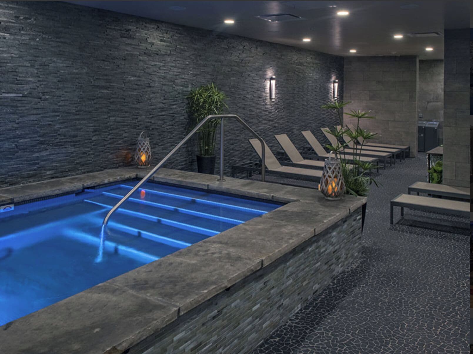 Un espace piscine intérieur moderne, agrémenté d’un mur en pierre, de chaises longues et d’un éclairage d’ambiance.