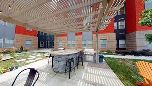 Un espace extérieur aménagé en terrasse, avec un bar en pierre et des sièges, entouré d’un bâtiment moderne aux murs rouges et gris.