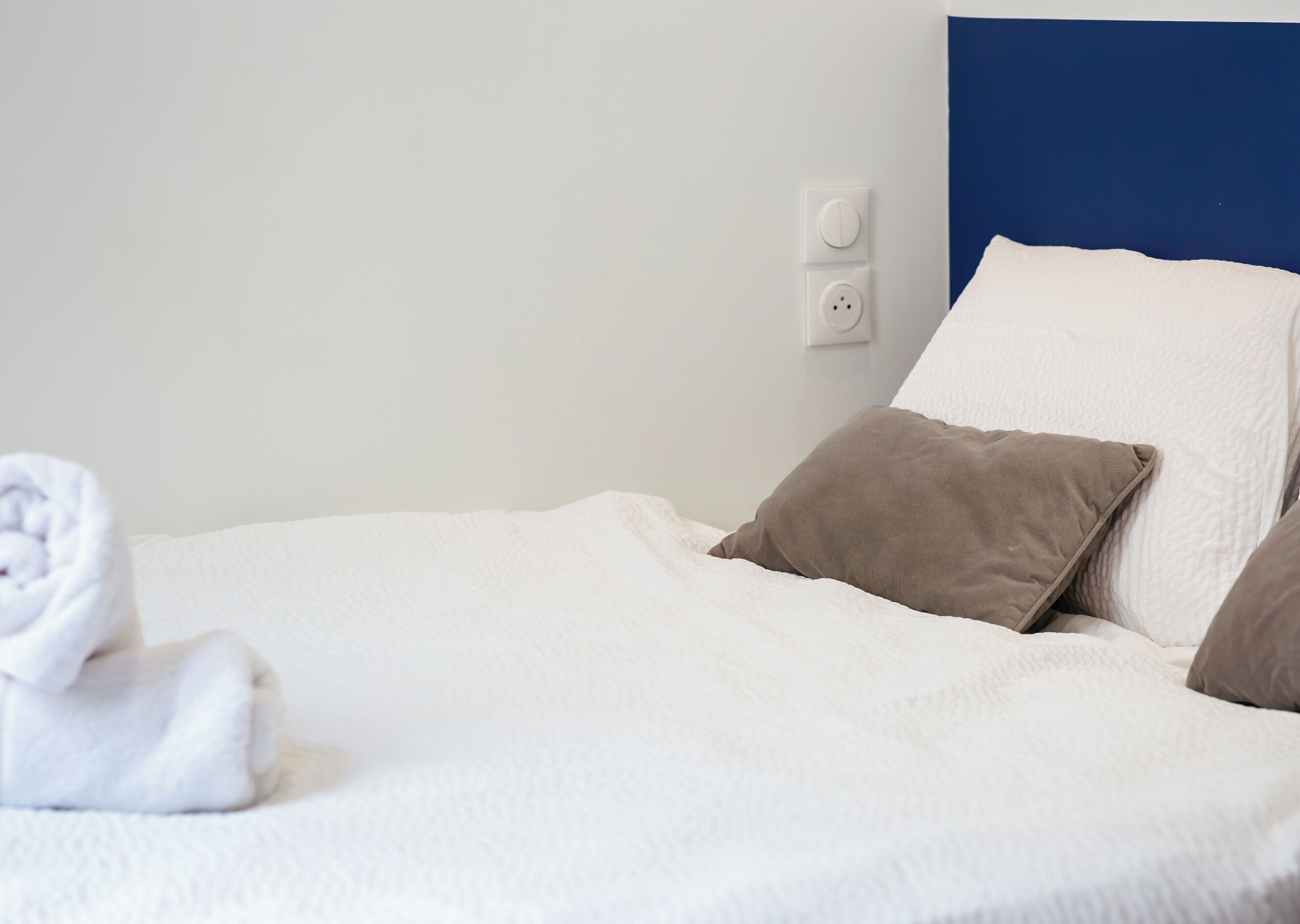 Ein ordentlich bezogenes Bett mit weißen und braunen Kissen, ein aufgerolltes weißes Handtuch und ein blauer Kopfteil.