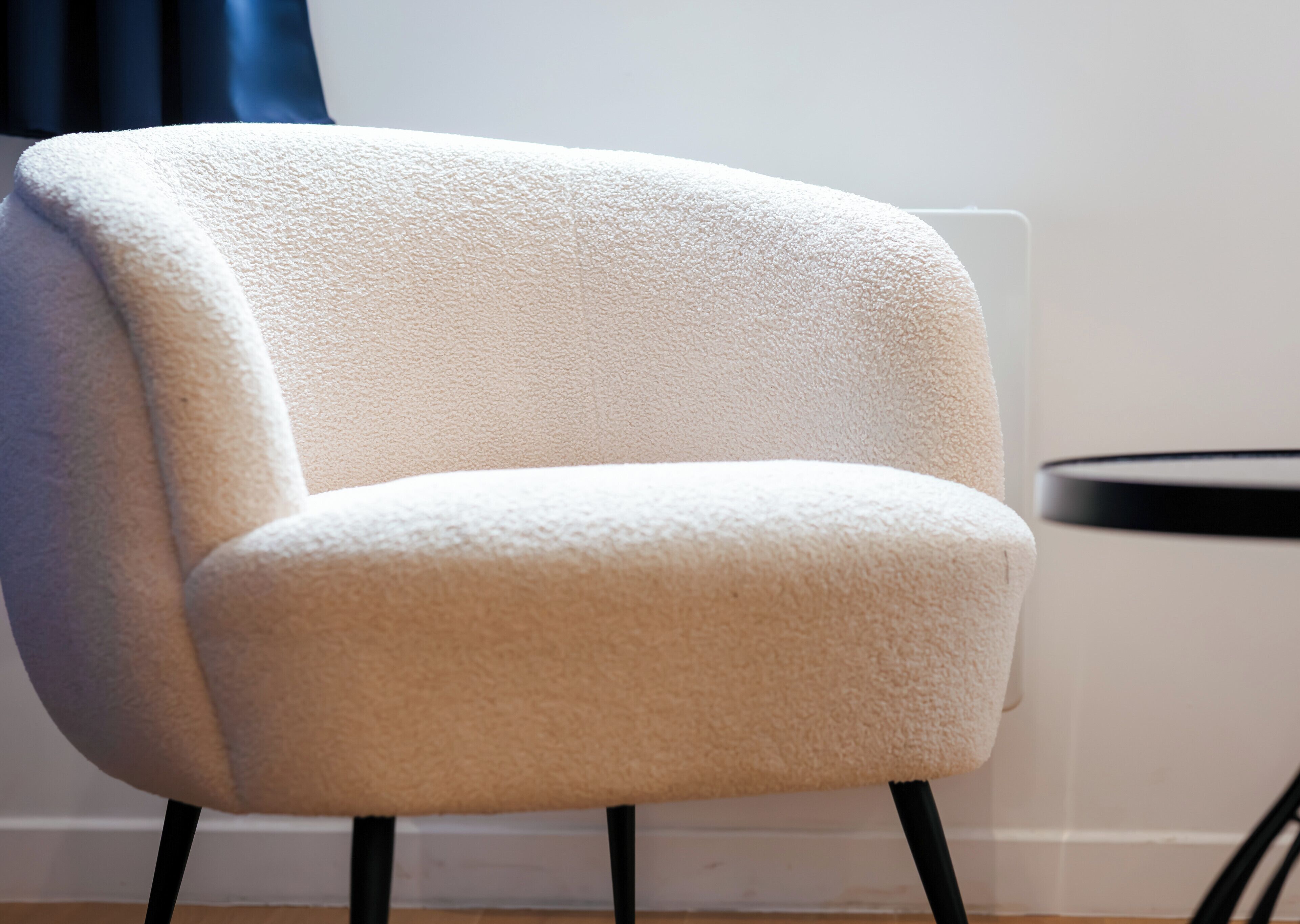 A white armchair with a textured fabric, positioned against a wall with a radiator and a dark curtain.