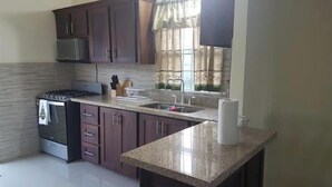 A kitchen with dark wooden cabinets, a stainless steel stove, a granite countertop, and a window with curtains. - Spacious two bedroom home with ocean view, pool and tennis court. (Florence Hall Village)