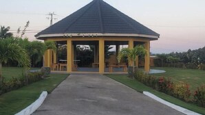 A gazebo-style structure with a dark roof and yellow walls, surrounded by a well-maintained lawn and palm trees. - Spacious two bedroom home with ocean view, pool and tennis court. (Florence Hall Village)