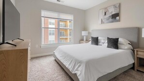 A bedroom with a large bed, a TV, a window with blinds, a painting, and a view of a building outside. - Landing | Stunning 2BD, Courtyard, Pool (Hyattsville)
