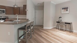 Cocina moderna con encimeras de granito, electrodomésticos de acero inoxidable y armarios de madera. Cuenta con un comedor con una mesa pequeña y sillas. El piso es de madera y las paredes están pintadas en un tono neutro.