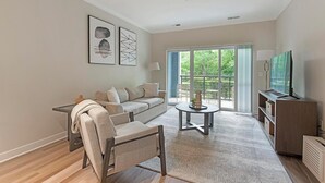 En moderne stue med sofa, sofabord og fjernsyn. Der er en balkon med udsigt til grønne omgivelser.