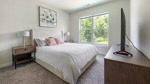 A bedroom with a large bed, two bedside tables with lamps, a TV on a stand, and a window with a view of trees.