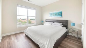 Een slaapkamer met een groot raam, een bed met witte lakens, een grijs hoofdeinde en een schilderij aan de muur.