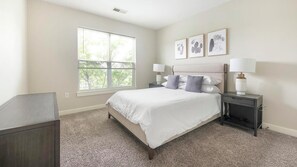 A bedroom with a large window, a bed with white bedding, two bedside tables with lamps, and three framed pictures on the wall.
