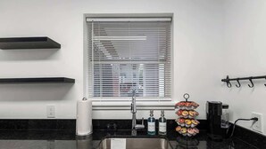 A modern kitchen with a stainless steel sink, black countertops, a window with blinds, and a rack with colorful bowls. - Modern UF Apartment ON Archer Rd Near Shands & VA (Gainesville)