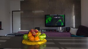 Un salon moderne avec un grand téléviseur à écran plat fixé au mur, un panier de fruits sur la table et un canapé agrémenté d’un coussin.