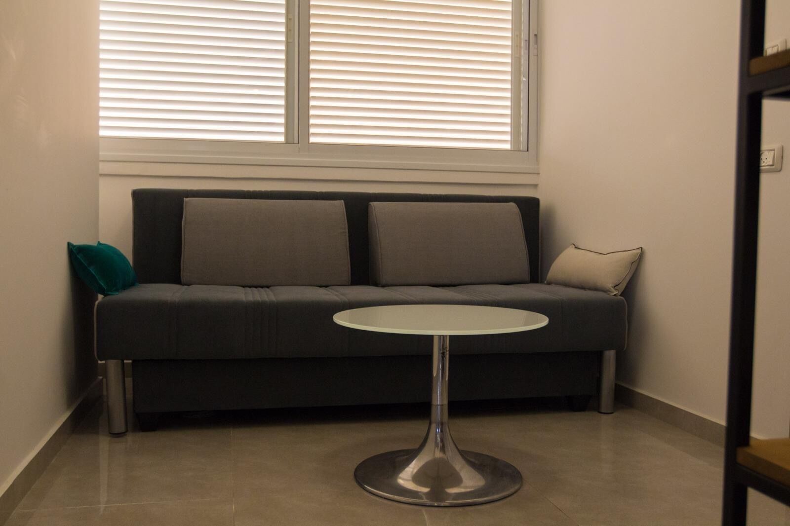 A dark grey sofa with two cushions, a small round table, and a window with blinds.
