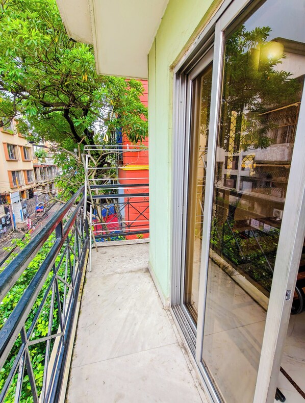 A balcony with a metal railing, a glass door, and a view of a tree and buildings in the background.