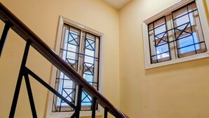 Una escalera con barandillas de madera y una pared amarilla. Se ven dos ventanas con barrotes de metal y marcos blancos.