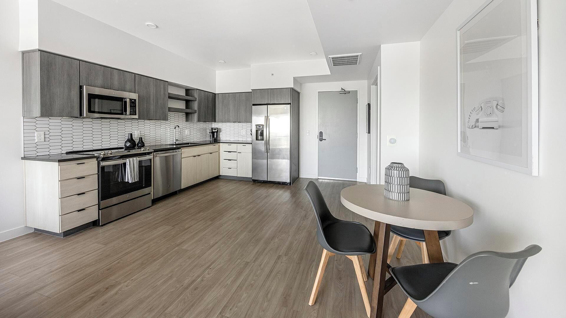 A modern kitchen with stainless steel appliances, a dining area with a round table and chairs, and a hallway leading to a door.