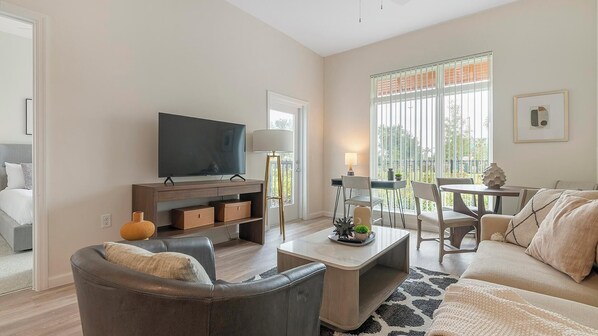A modern living room with a flat-screen TV, a sofa, a coffee table, and a dining area with chairs and a table. - Landing | Beautiful 2BD, Game Room, Pool (Orlando)