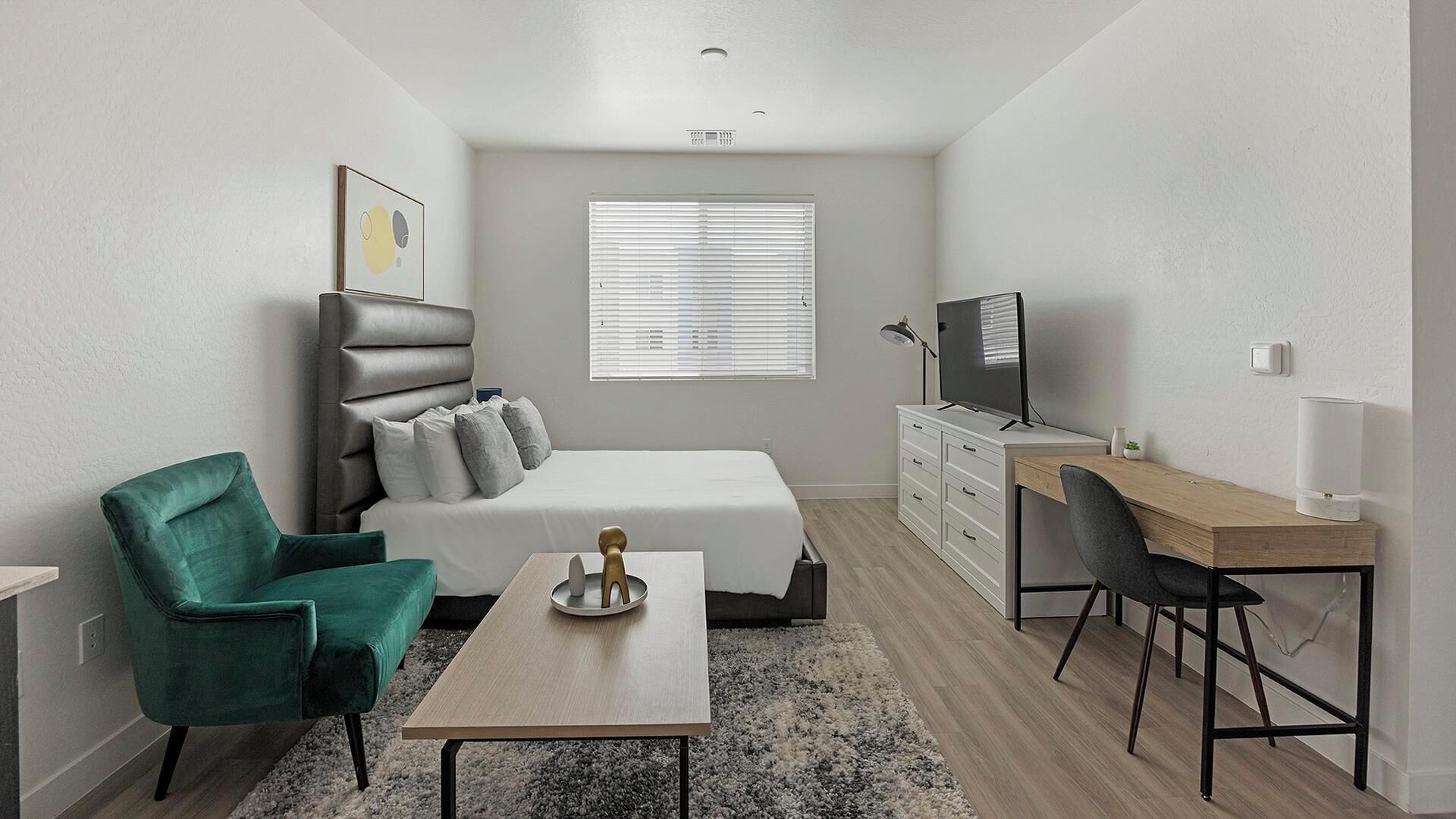 Une chambre à coucher de taille réduite, comprenant un lit, un bureau, une chaise, un téléviseur et une fenêtre avec des stores.