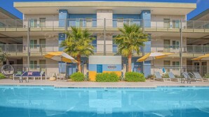 A multi-story hotel with a pool area, palm trees, and a caution sign for shallow water.