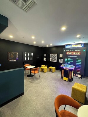 Een kamer met een Pac-Man spelcomputer, een tafel met stoelen en een muur waarop de hoogste scores staan.
