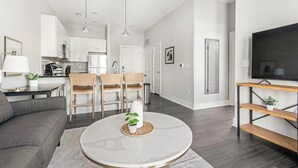 Un salon moderne avec un canapé gris, une table basse ronde ornée d’une plante et un téléviseur posé sur un meuble en bois.