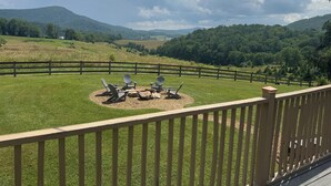 A grassy outdoor area with Adirondack chairs and a fire pit, surrounded by a wooden fence, overlooking a scenic landscape with hills and trees.