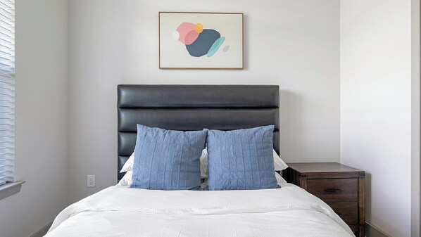 A bedroom with a bed, a dark headboard, two blue pillows, a white comforter, a wooden nightstand, and a wall-mounted abstract painting.