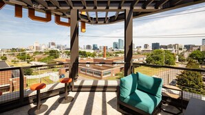 Un balcón con vistas a un paisaje urbano, una silla color turquesa y dos taburetes naranjas.
