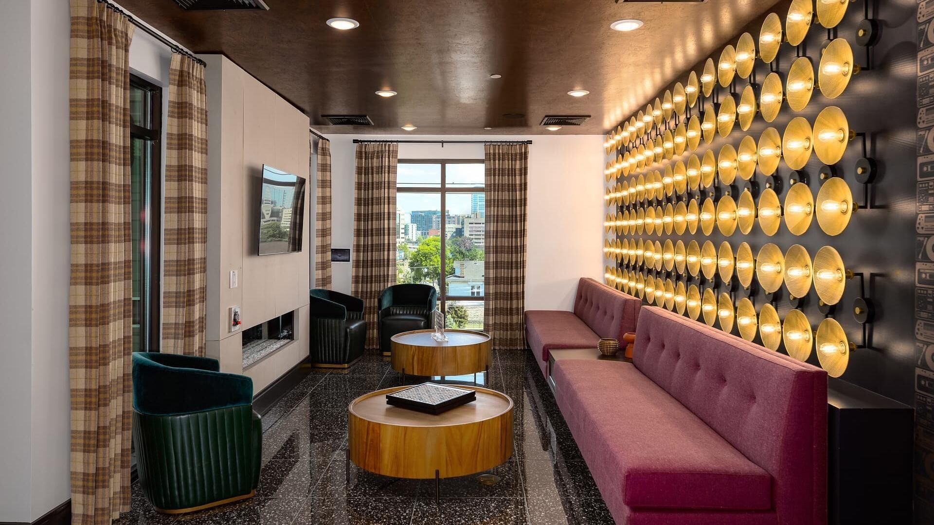 A modern hotel lobby with a large window, a round wooden table, a plush pink sofa, and a wall of decorative lights.