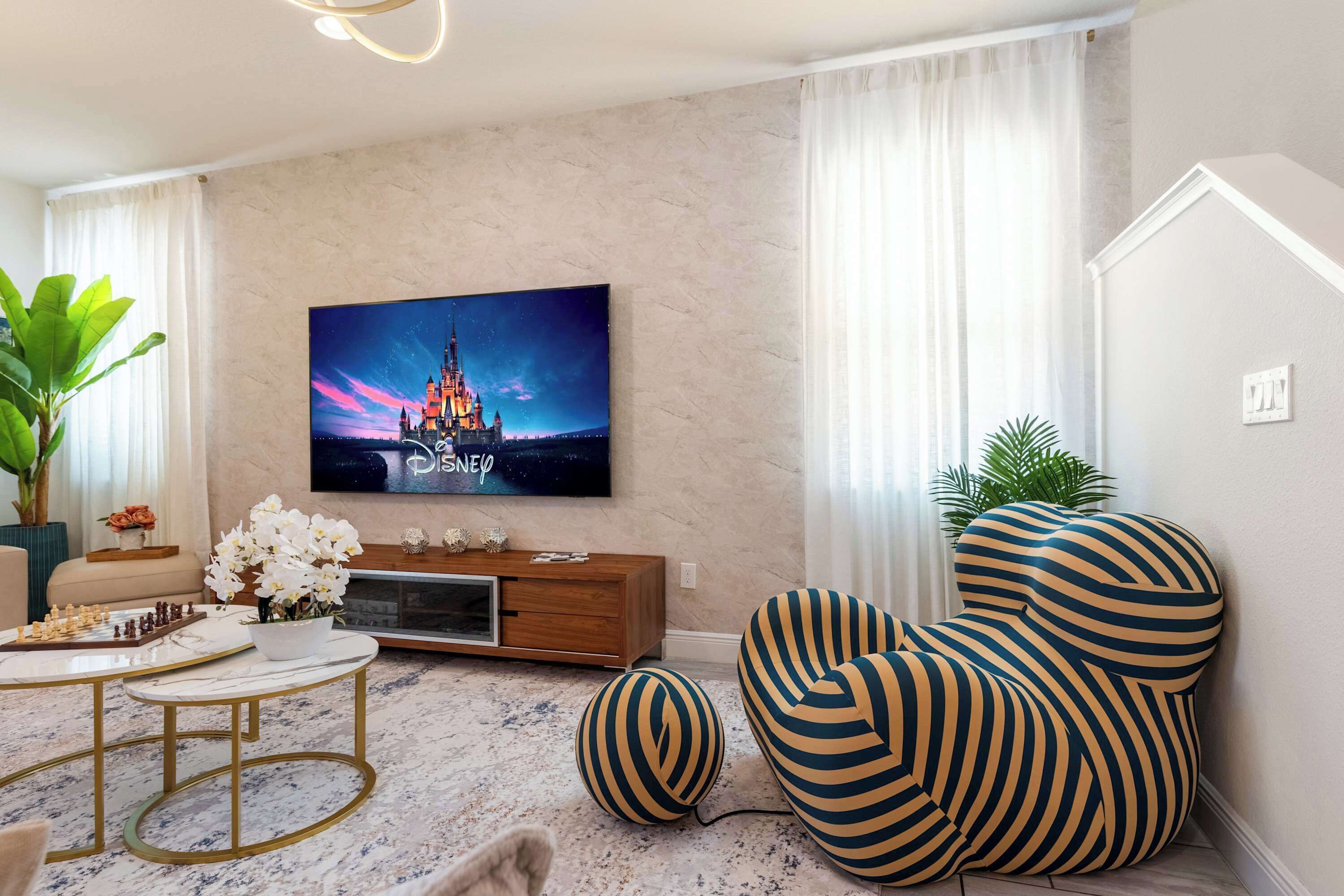 Ein modernes Wohnzimmer mit einem Flachbildfernseher, einem Marmortisch, gestreiften Sesseln und einer großen Topfpflanze.