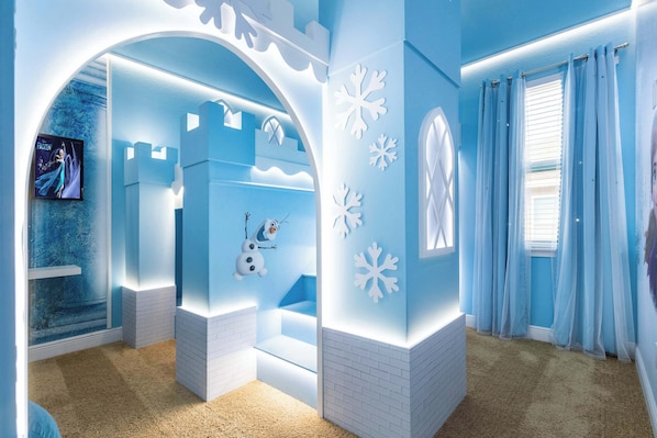 Une pièce aux couleurs bleu et blanc, décorée avec un bonhomme de neige, équipée d’un écran de télévision et dotée d’une fenêtre avec des rideaux.