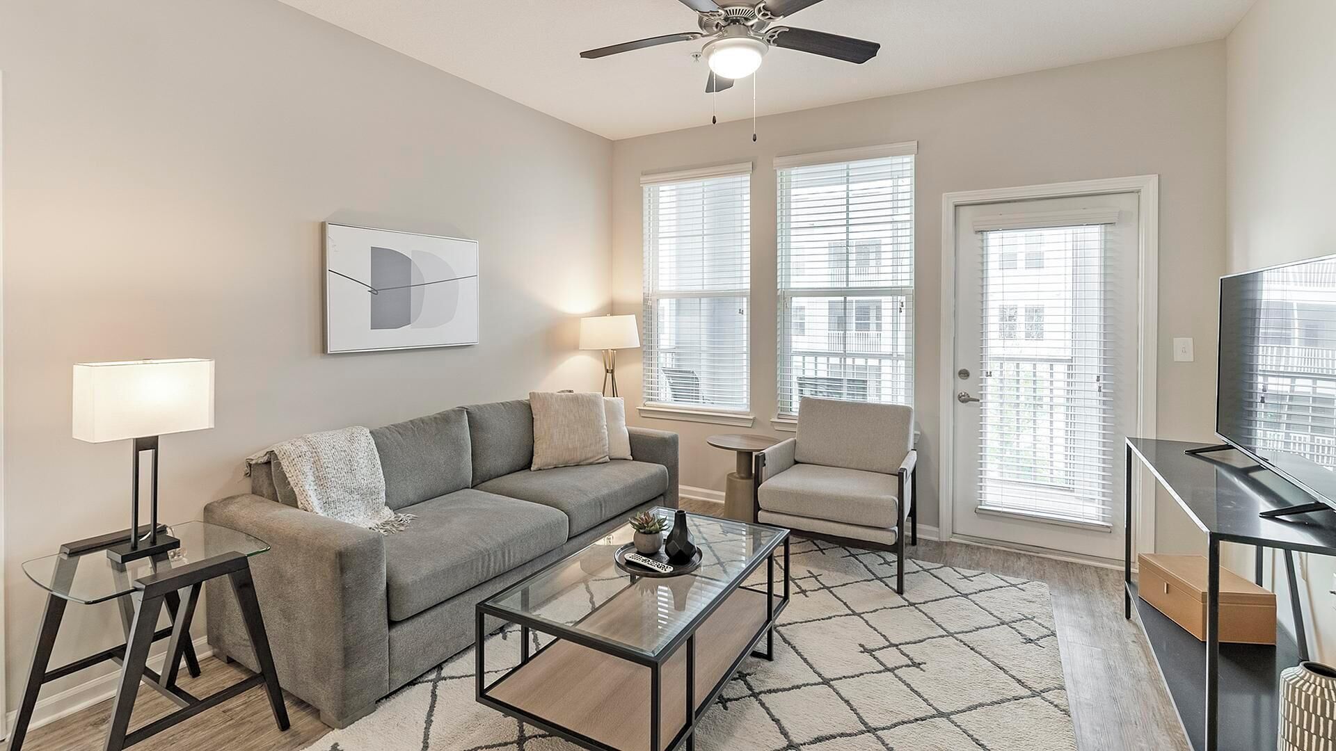 A modern living room with a grey sofa, a glass coffee table, a side table with a lamp, a floor lamp, and a ceiling fan.