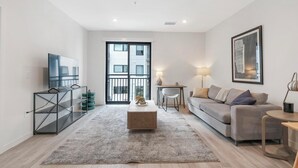 Een moderne woonkamer met een flatscreen televisie, een grijze bank, een salontafel en een groot raam met een balkon.