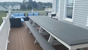 A wooden deck with a long table and benches, white railings, and a view of a house and trees in the background. - Amazing 10 Bedroom Compound on the Water with 6 Bedroom Home & 4 Studio Cottages (Stonington)