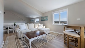 A spacious living room with a white sectional sofa, a wooden coffee table, and a dining area with a wooden table and chairs. A large window offers a view of the outdoors. - Amazing 10 Bedroom Compound on the Water with 6 Bedroom Home & 4 Studio Cottages (Stonington)
